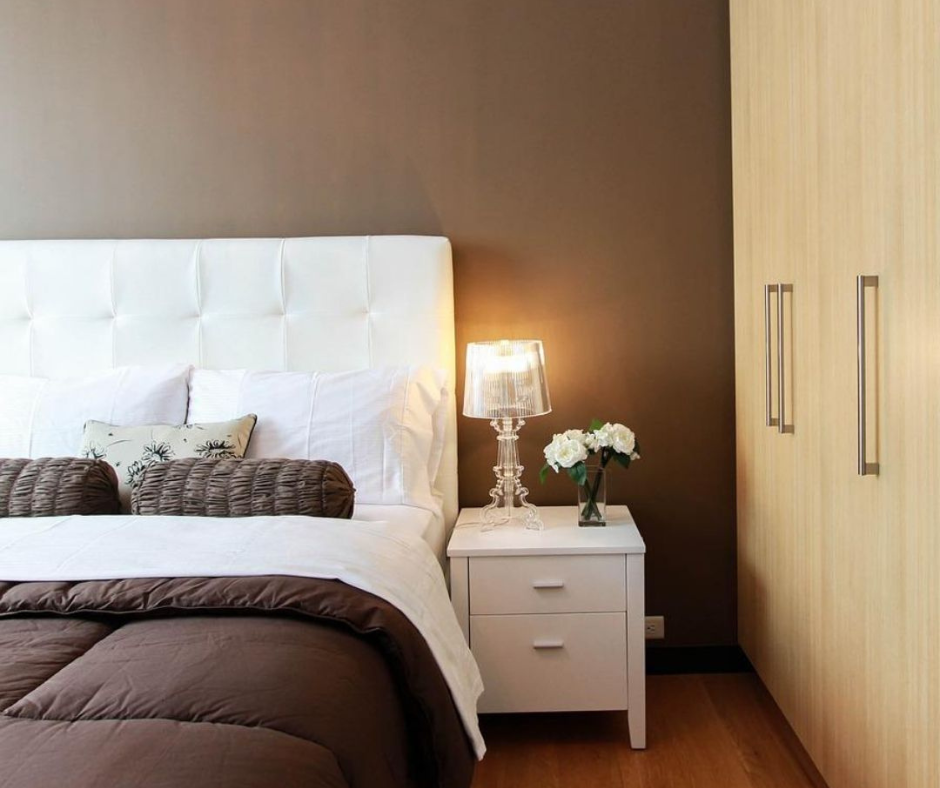 Picture of one side of a bed with a white headboard and brown comforter. The wall behind the bed is painted brown. There is a white nightstand beside the the bed and a wood closet to the right of the nightstand.