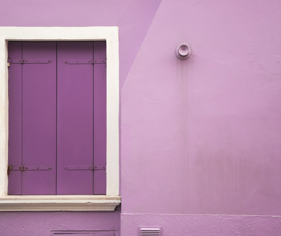 Picture of a window shutter painted purple framed by white trim inside a lavender painted wall.