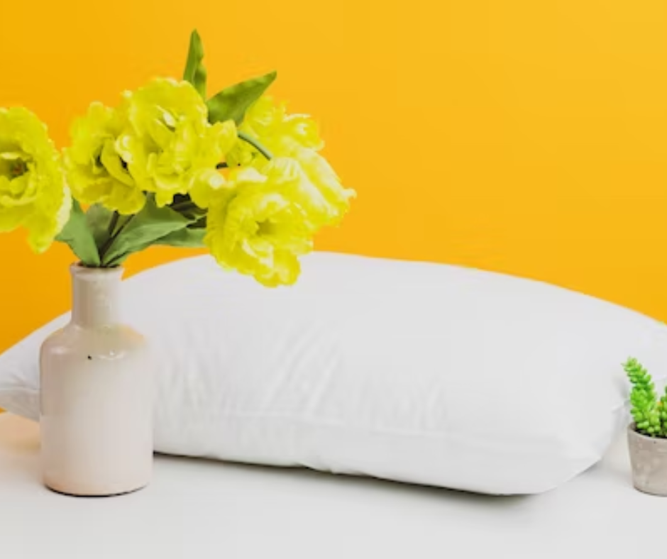Picture of a white pillow sitting beside a white vase with yellow flowers in it. The wall behind is bright yellow.